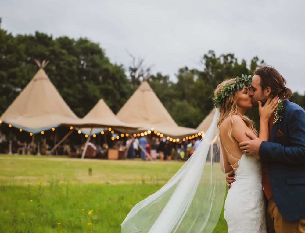 A romantic wedding couple kisses in front of tipi tents during an outdoor festival-style celebration in a green field.