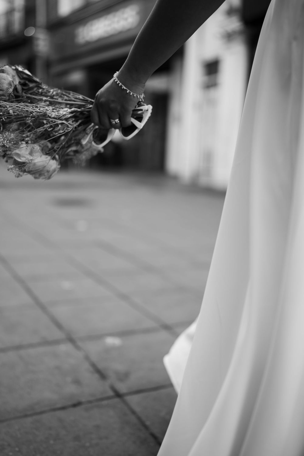 Black and white photo of a bride holding a bouquet and heels while wearing a flowing wedding dress on a city sidewalk.