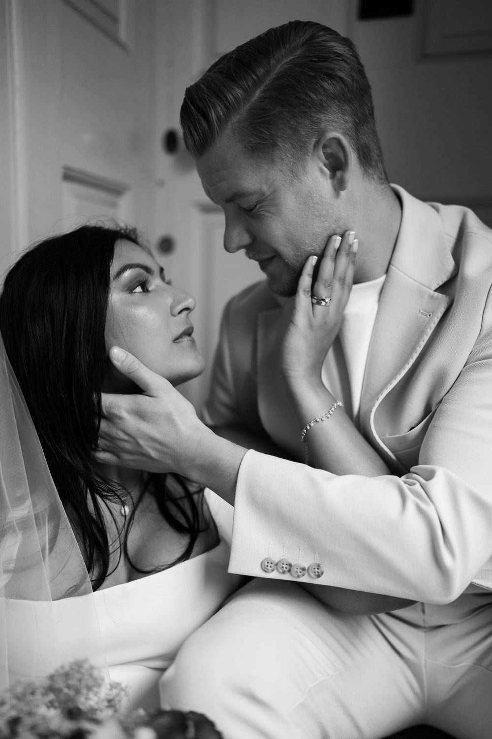 Black and white wedding photo of bride and groom gazing at each other tenderly, bride wearing veil, groom in white suit.