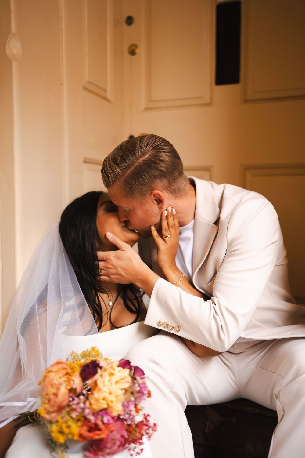 A bride and groom share a romantic kiss, she in a white gown with veil holding a colorful bouquet, he in a white suit.