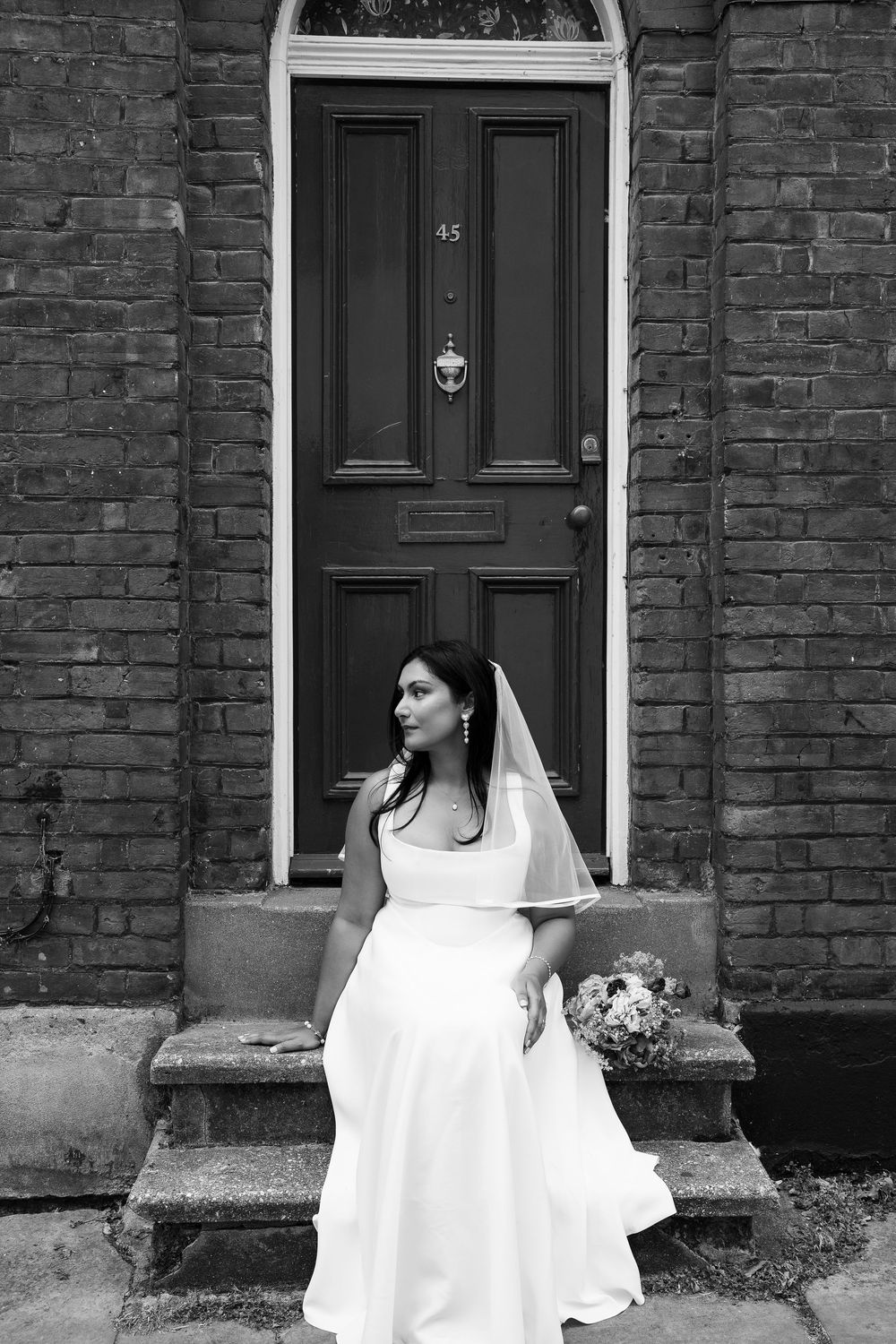 Black and white photo of a bride in a white gown and veil holding a bouquet on steps outside a brick building.