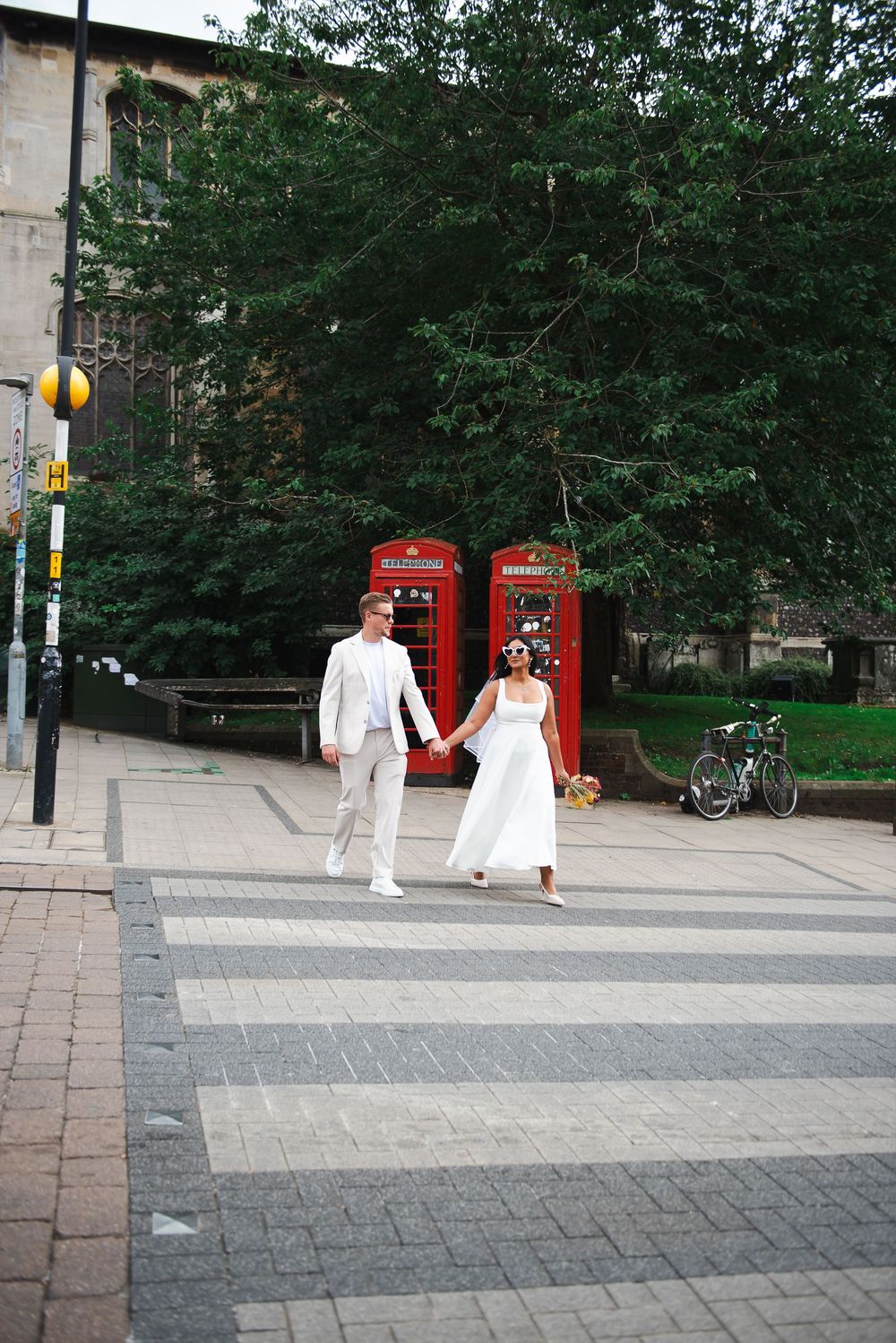 A couple in wedding attire holds hands crossing a striped plaza with iconic red London phone boxes behind them.