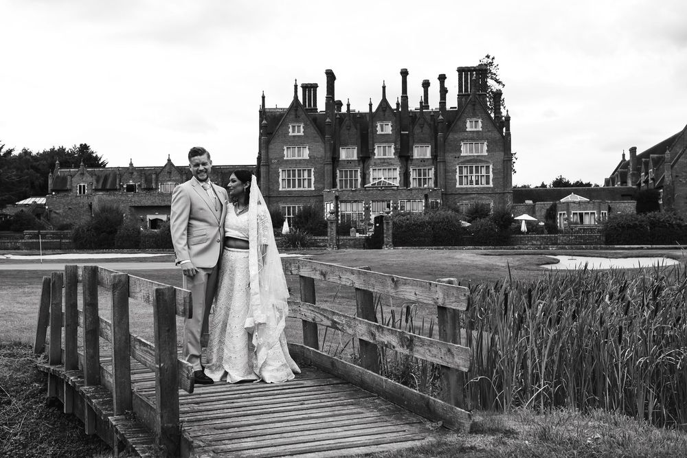 Black and white photo of a bride and groom posing on a wooden bridge outside a grand historic manor house.