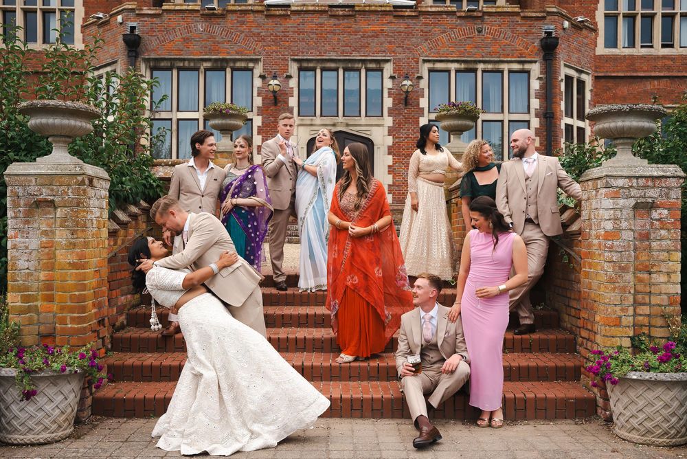 Wedding party poses on brick building steps, bride and groom share a dip kiss while guests celebrate around them.