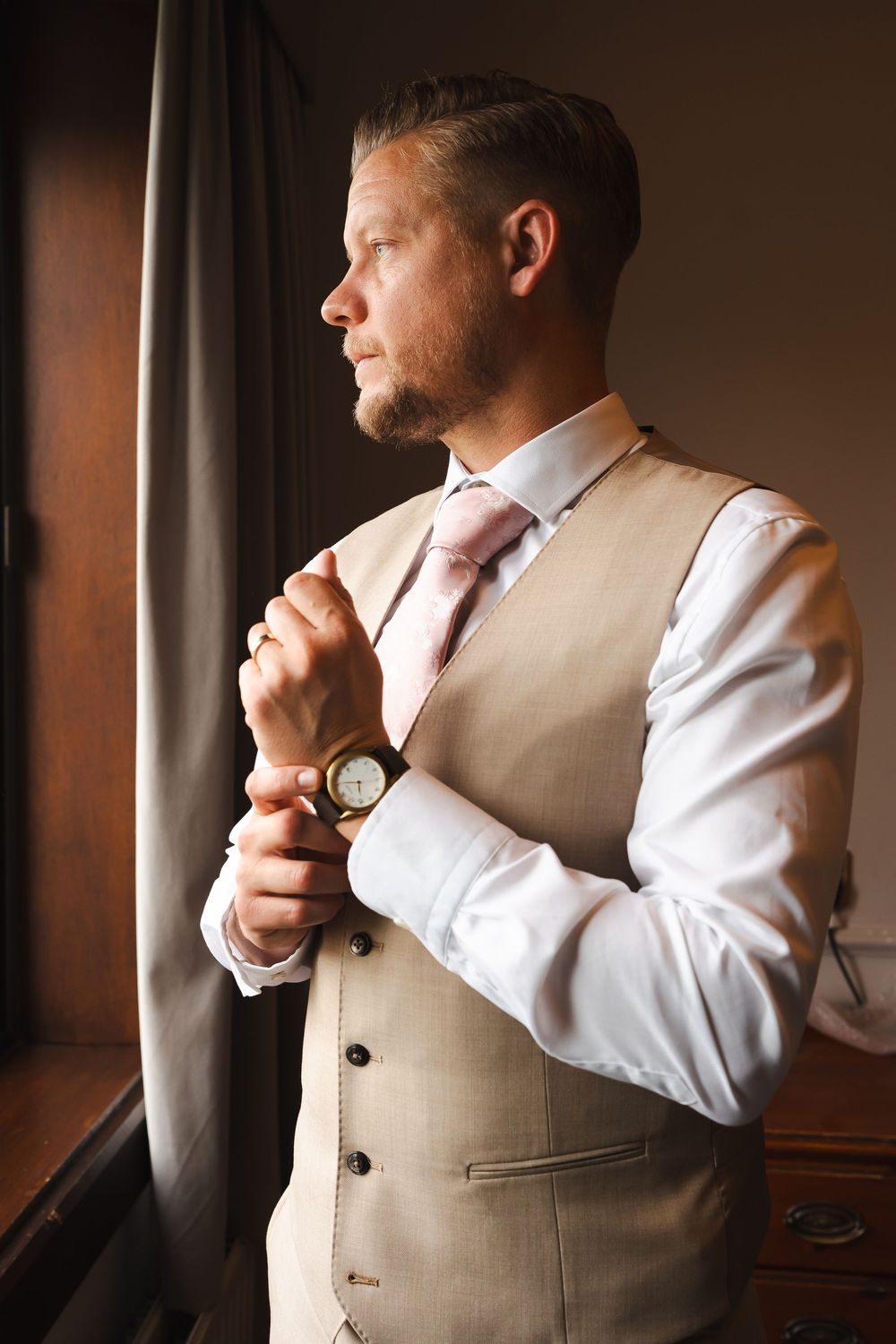 Elegant groom in beige vest and pink tie adjusting cufflinks by window, preparing for wedding ceremony.