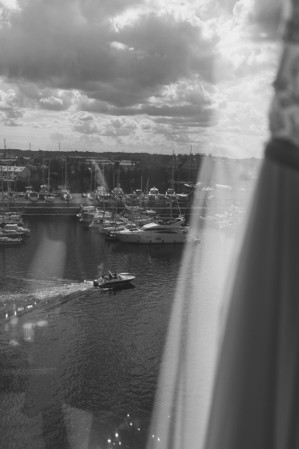 Black and white aerial view of a marina with boats on calm water, dramatic cloudy sky, shot through a window.