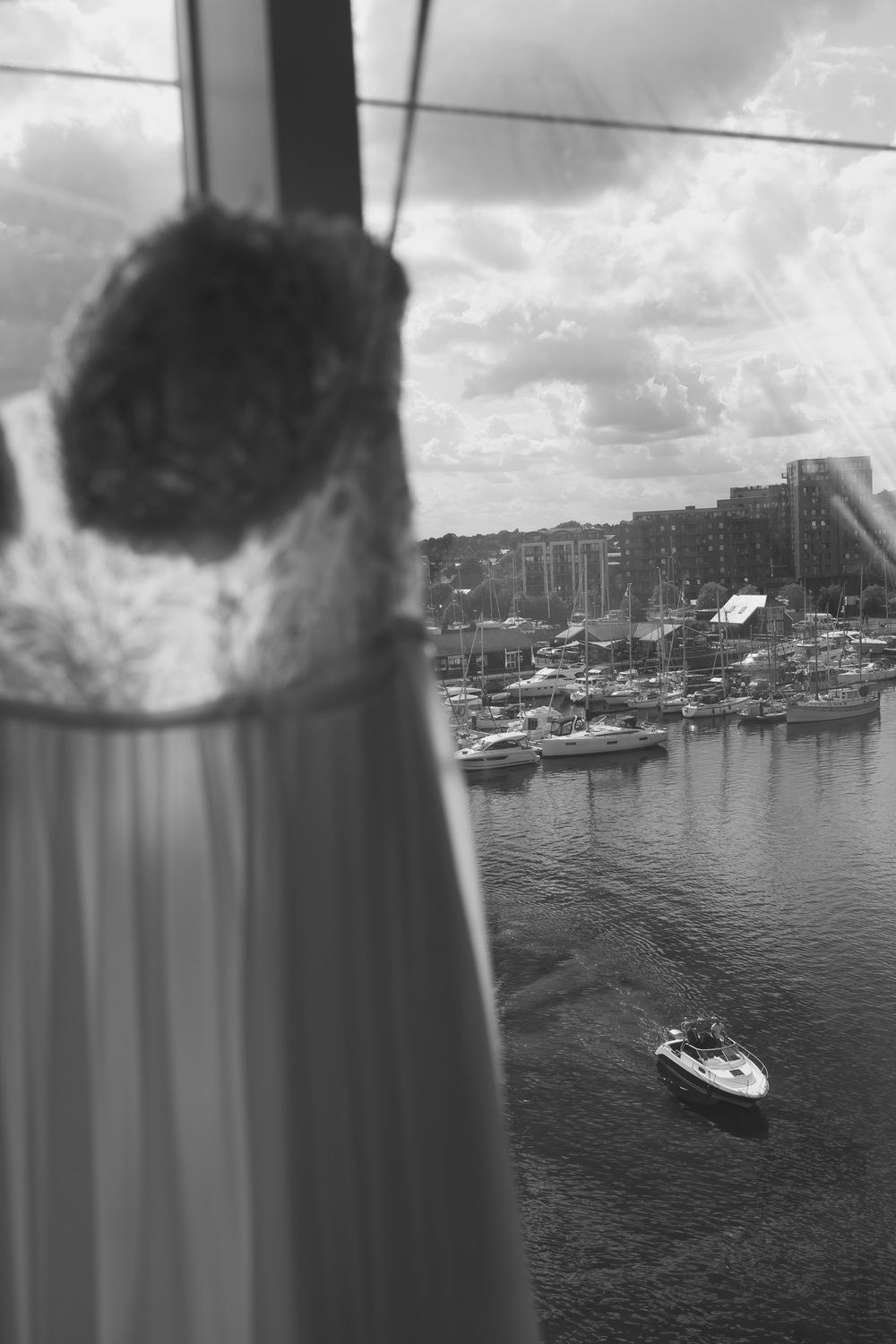 Black and white photo of a wedding dress hanging by a marina window with boats and cloudy sky in background.