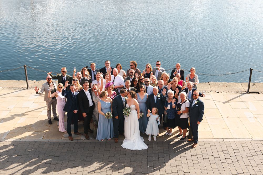 Large wedding group photo of bride, groom and guests posing on a sunny waterfront dock.