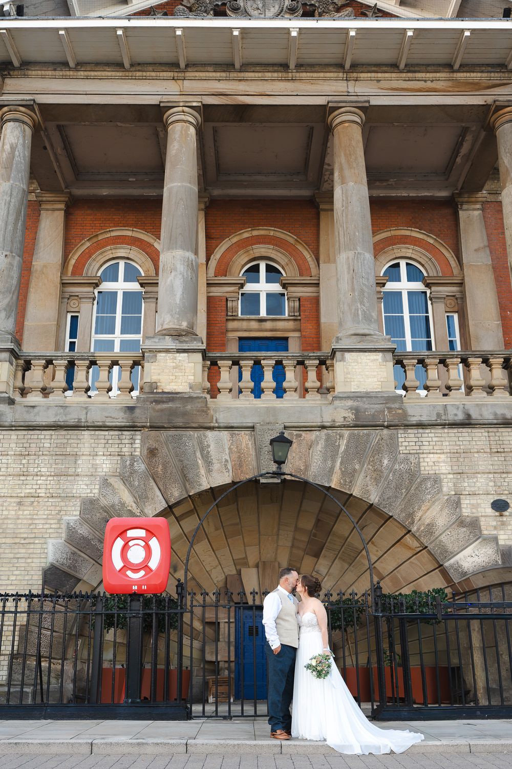 Bride and groom kissing outside a grand Victorian building with arched entrance, columns, and red brick facade.
