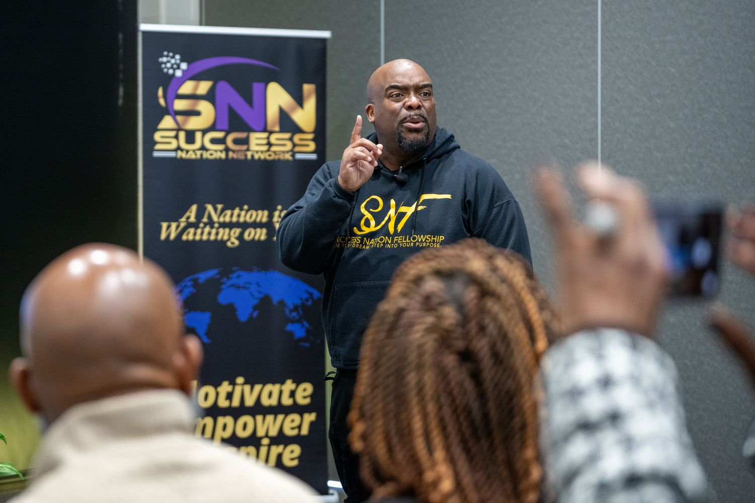 A speaker gestures while presenting at a motivational business conference with banners visible in the background.
