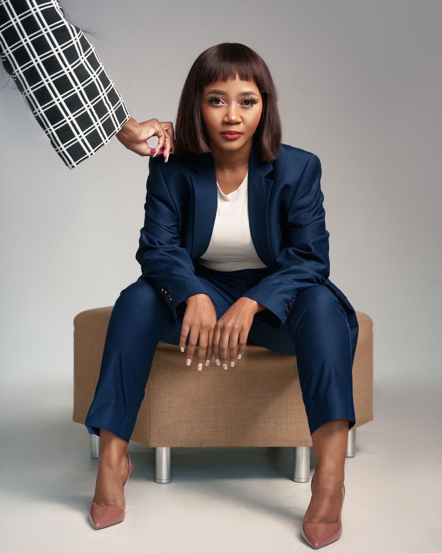 Professional in a navy blue suit sits confidently on a beige chair holding a black and white prop against grey backdrop.