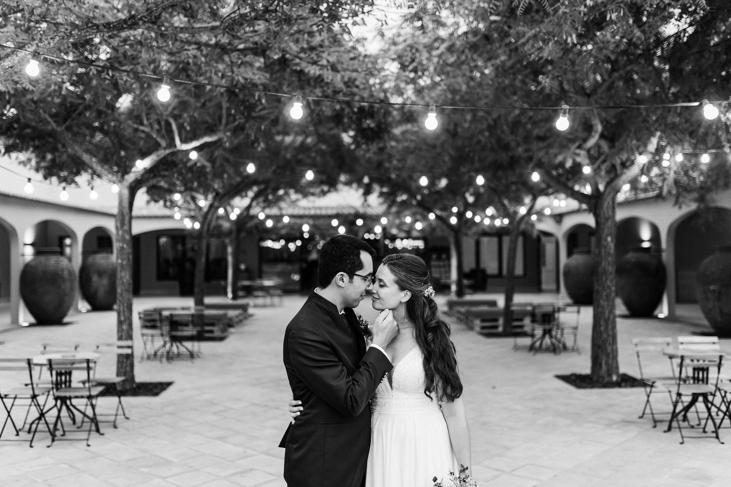A romantic black and white moment under outdoor string lights in a courtyard with bistro tables and trees.