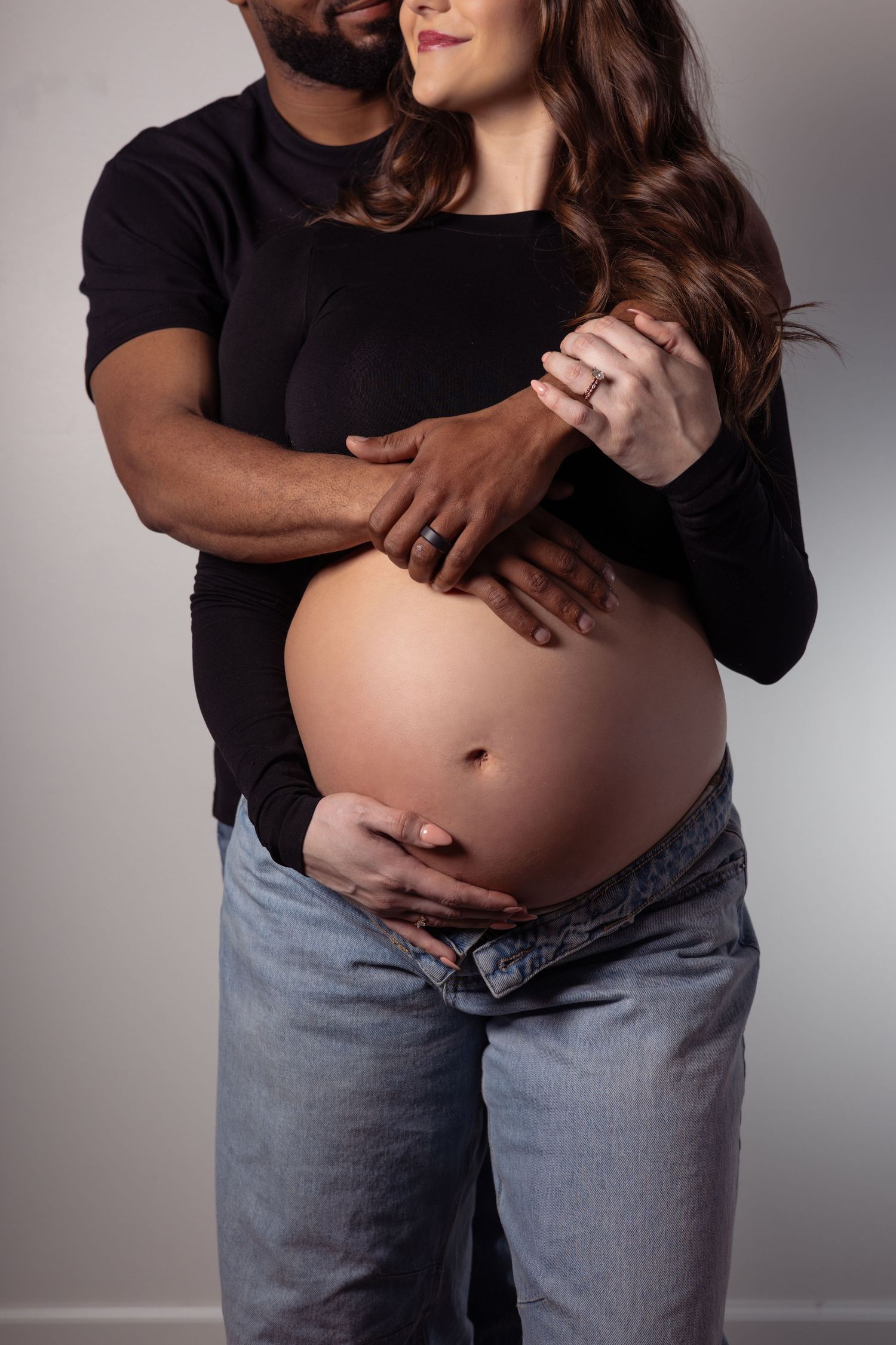 Pregnant woman in black top and jeans holds ultrasound images while someone in white touches her belly.