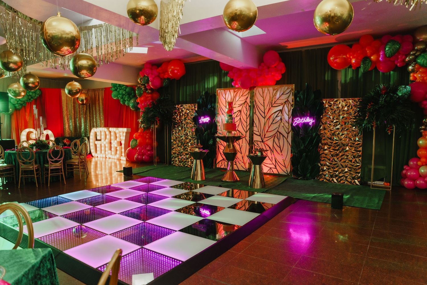 Dance floor with illuminated checkered tiles surrounded by gold disco balls, red flowers and green foliage in an event space.