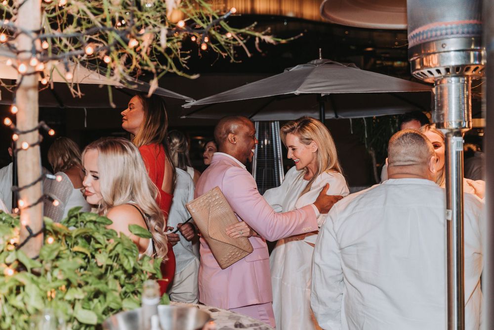 People gather under a patio umbrella at an outdoor event with decorative greenery and lighting at dusk.
