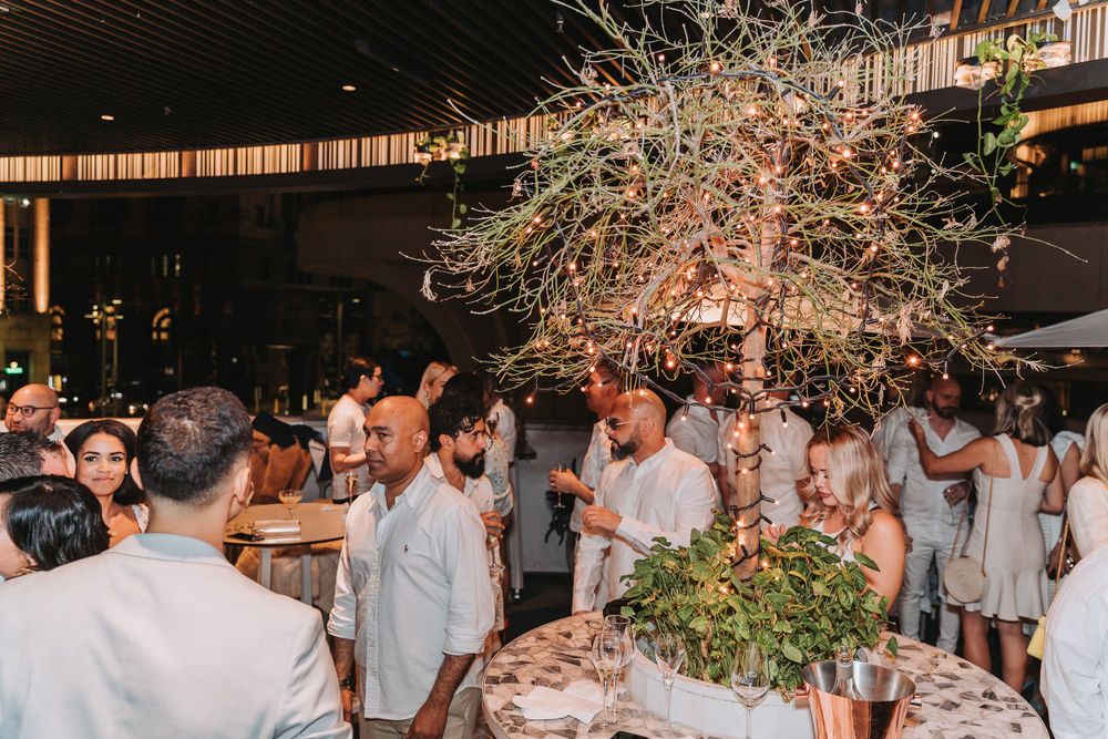 Guests in white attire gather at an elegant evening party with string lights and floral decorations.