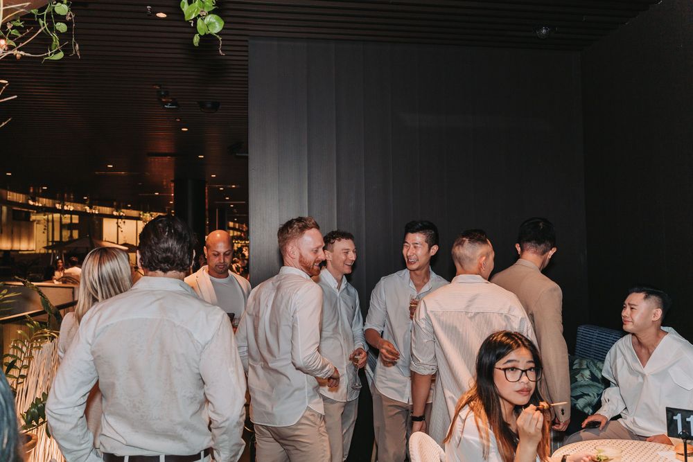 A group of people in light colored shirts gather for drinks at an evening social event in a dimly lit venue.
