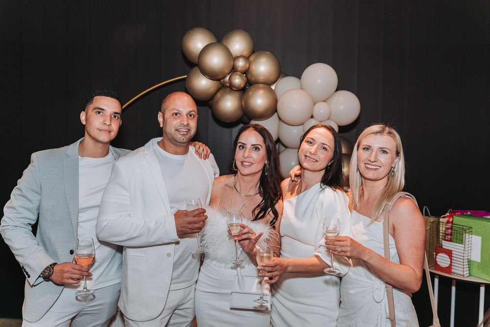 Group in white attire poses with drinks at party under metallic balloon arch decor against dark background.