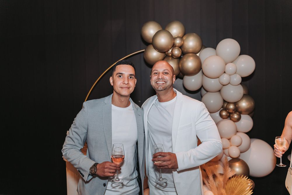 Two people in light colored outfits pose together at a party with gold and white balloon decorations in the background.