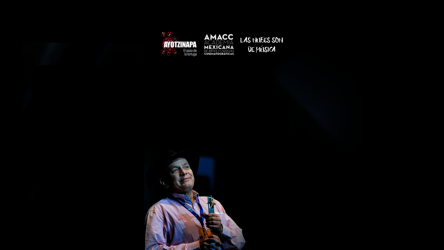 A performer speaks on stage at the AMACC Mexican cinema awards ceremony under dramatic dark lighting.