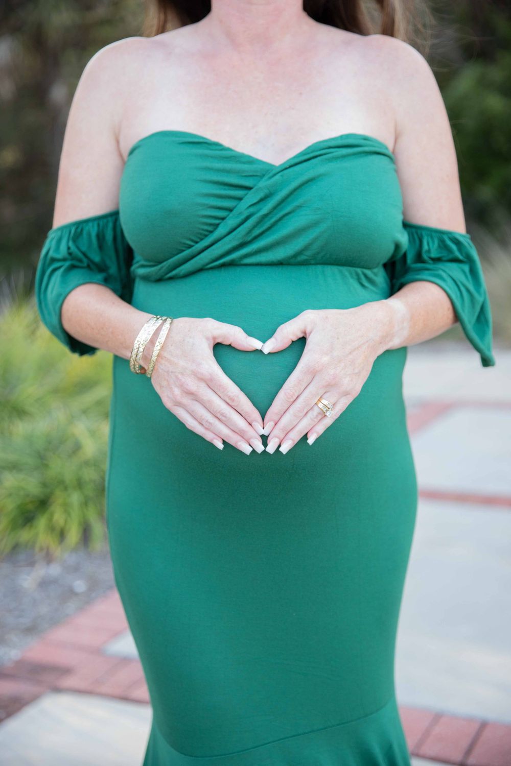 Pregnant woman in emerald green off-shoulder gown making heart shape with hands over baby bump.