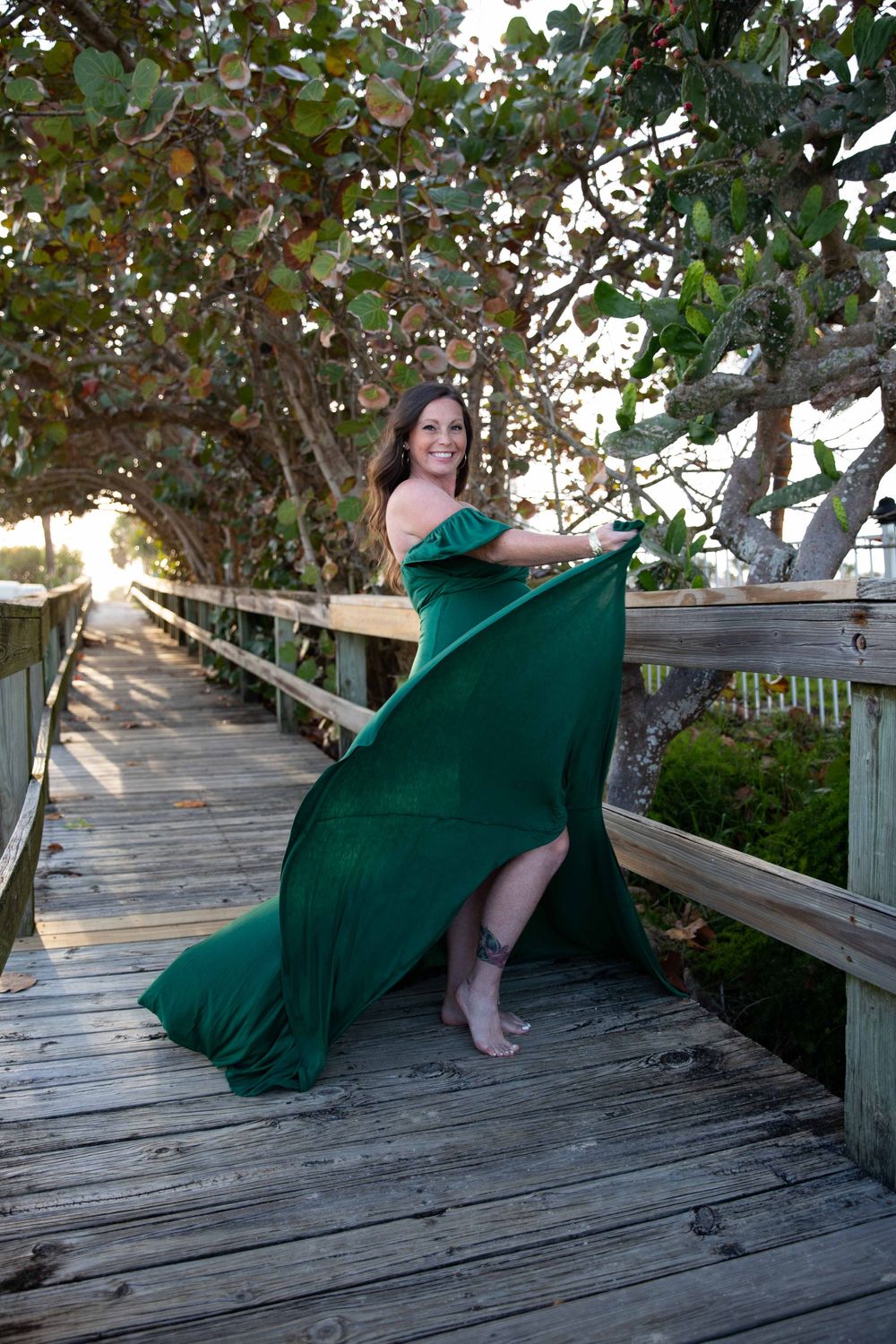 Woman in flowing emerald green off-shoulder gown twirling on wooden boardwalk surrounded by trees.
