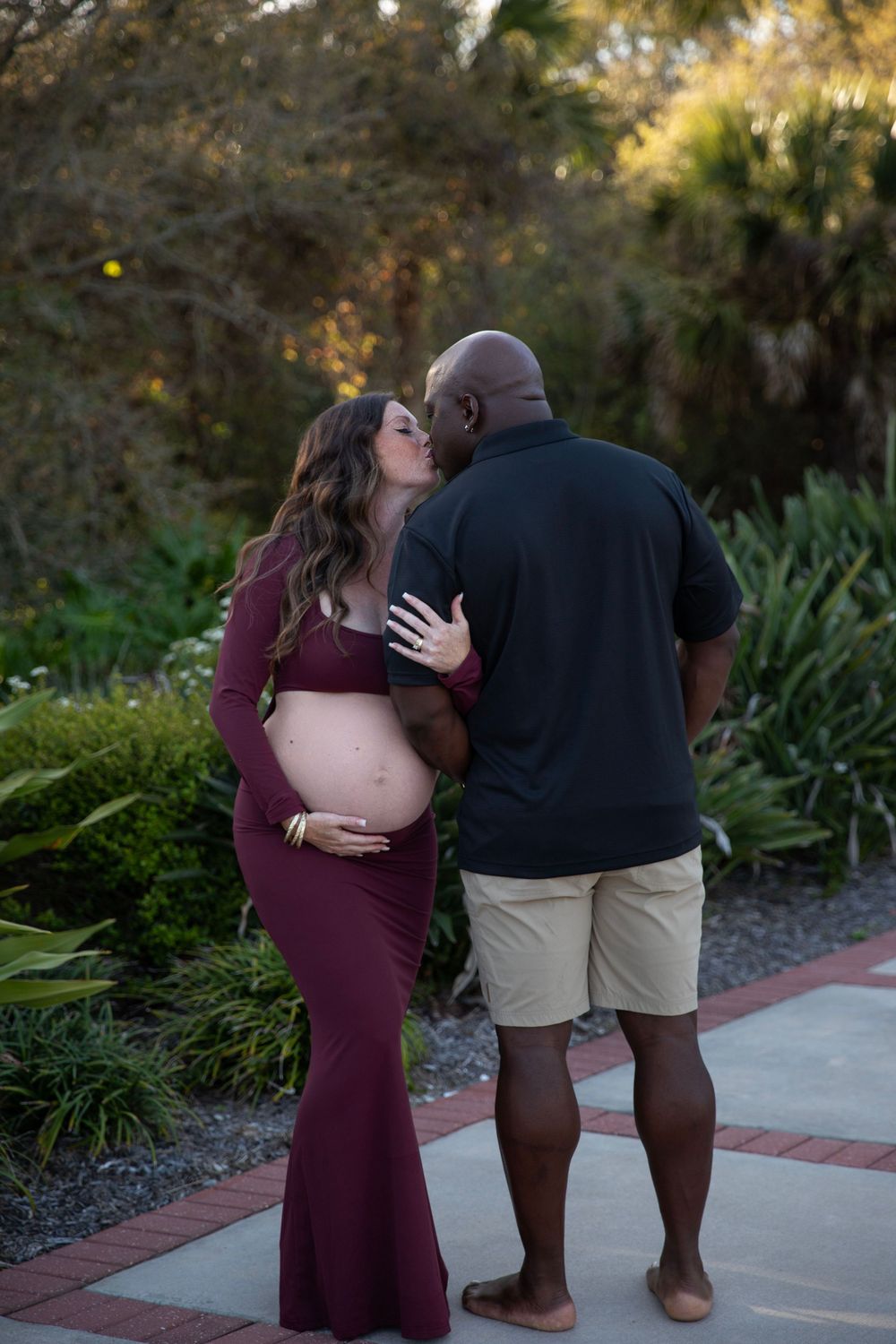 Pregnant couple sharing a kiss outdoors, woman in burgundy form-fitting maternity gown.