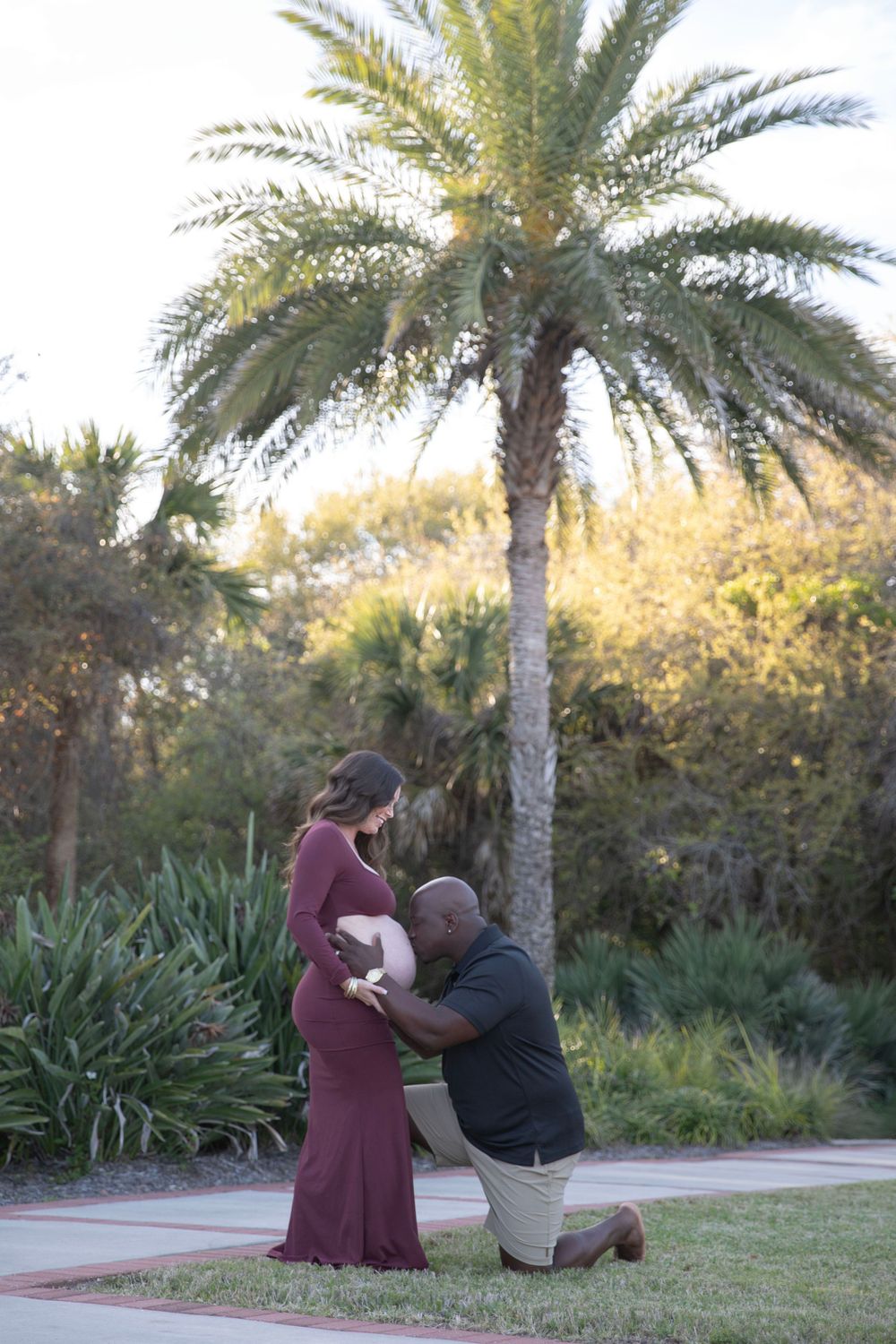 Man kneeling to kiss pregnant womans belly outdoors near palm tree during maternity photo session.