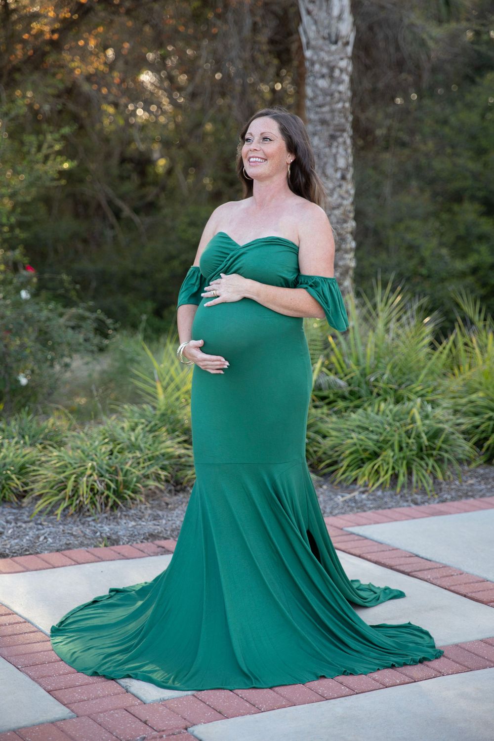 Pregnant woman in elegant emerald green off-shoulder mermaid maternity gown posing outdoors.