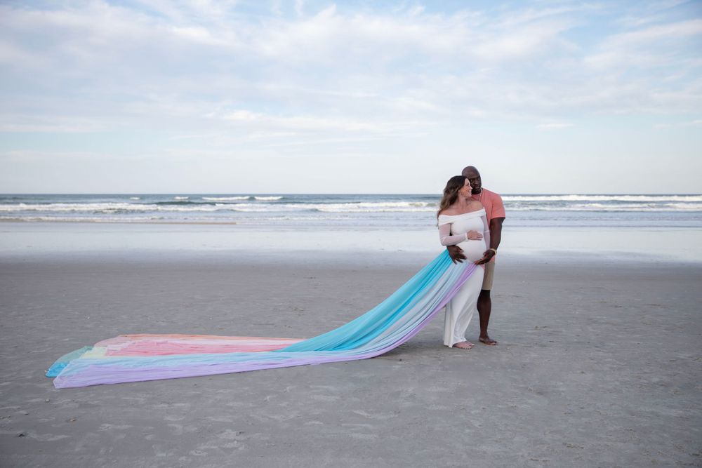 Couple kissing on beach with long pastel rainbow fabric flowing behind them during maternity shoot.