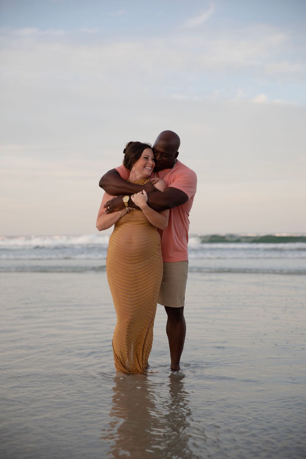 Interracial couple embracing on beach shore, pregnant woman in gold dress with partner in pink shirt.