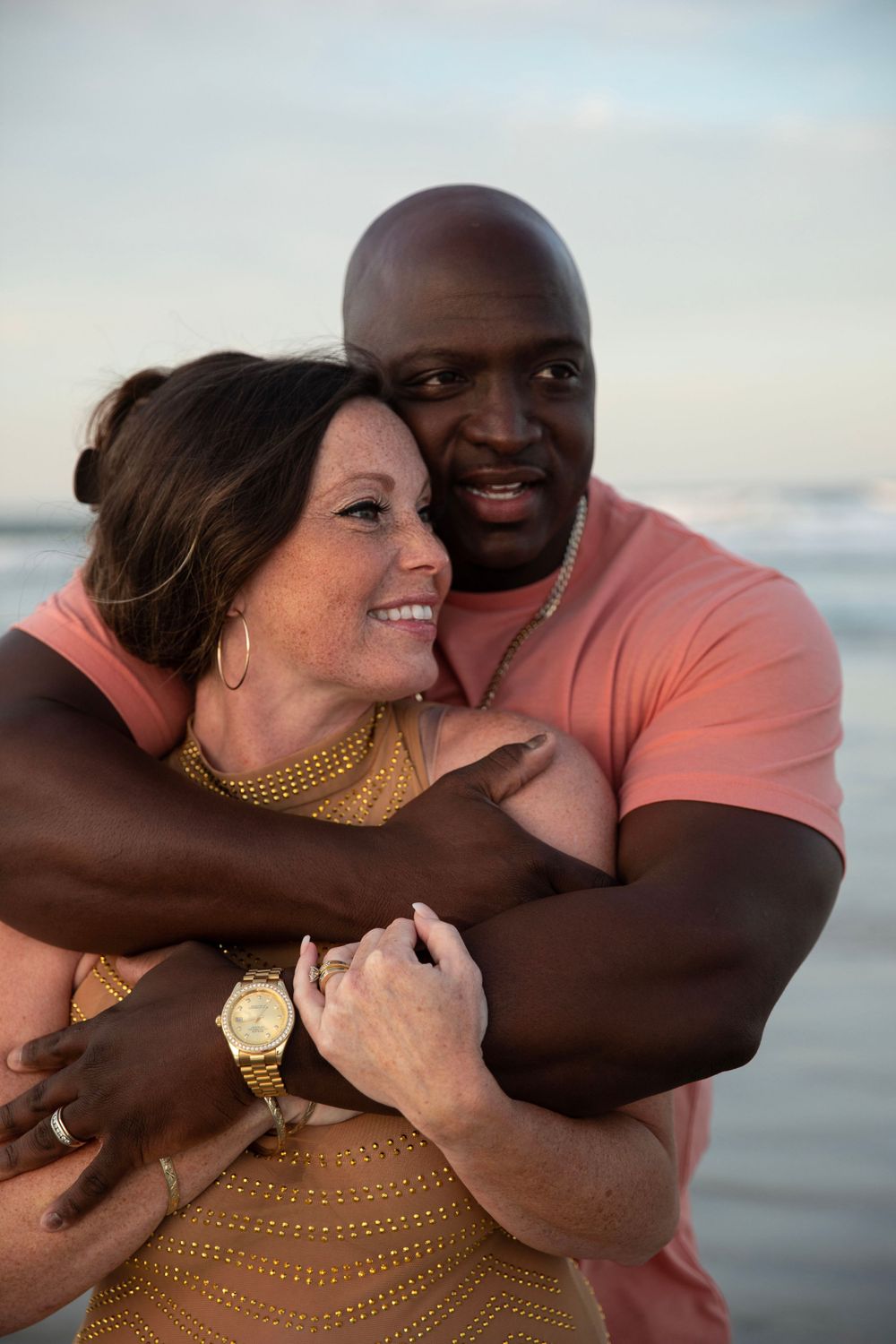 Close-up of interracial couple hugging on beach, man in pink shirt embracing smiling woman in gold dress.
