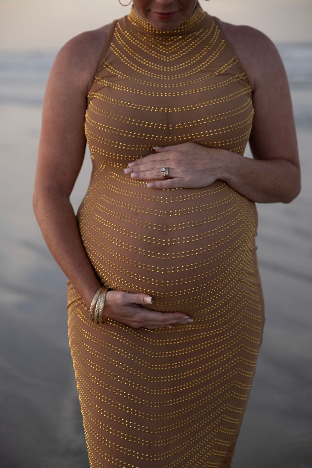 Close-up of pregnant woman in gold striped maternity dress cradling baby bump with both hands.