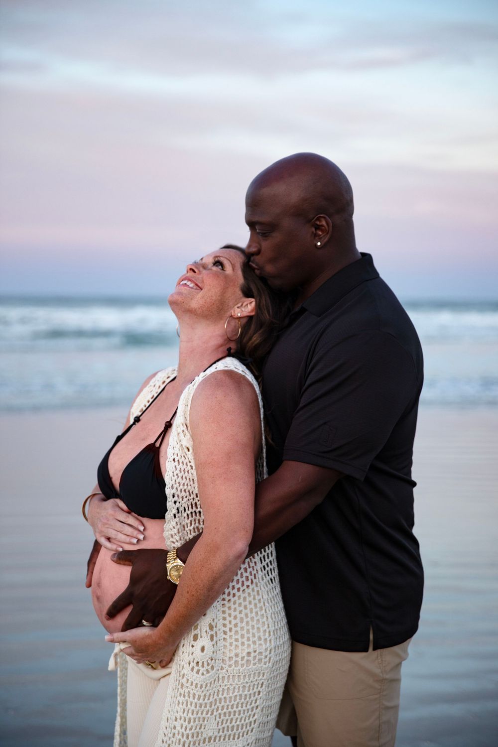 Interracial couple on beach at sunset, man kissing pregnant womans temple while holding her bump.