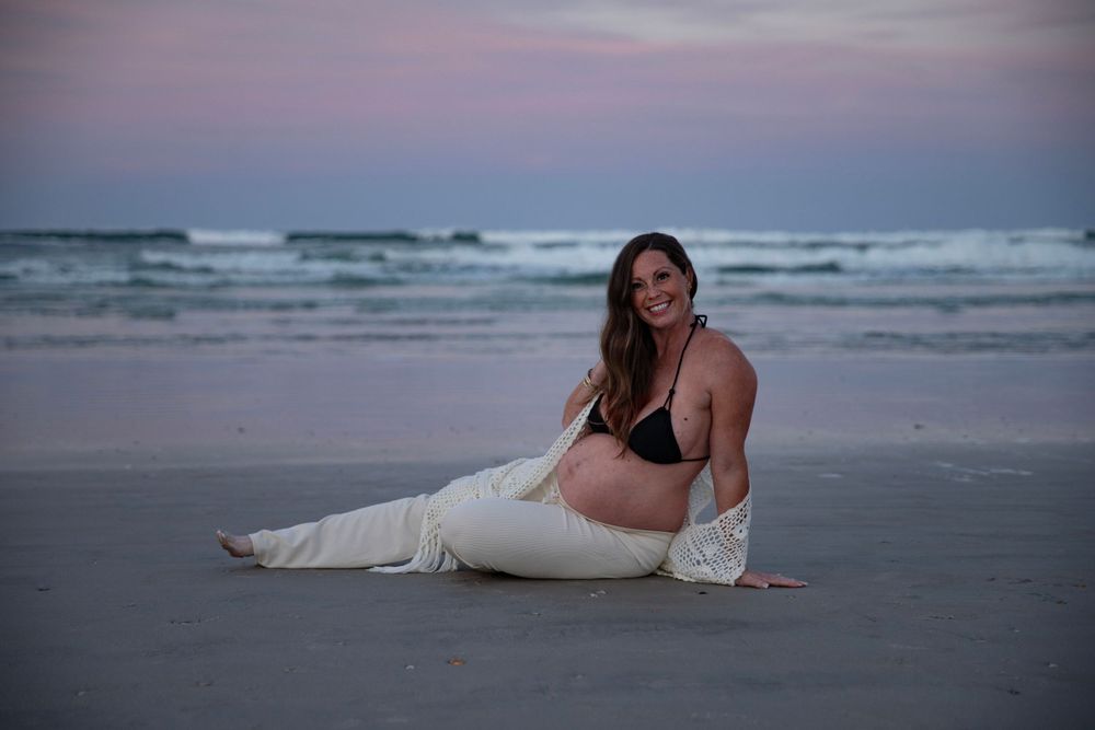 Pregnant woman in black bikini and white pants posing on sandy beach at dusk.
