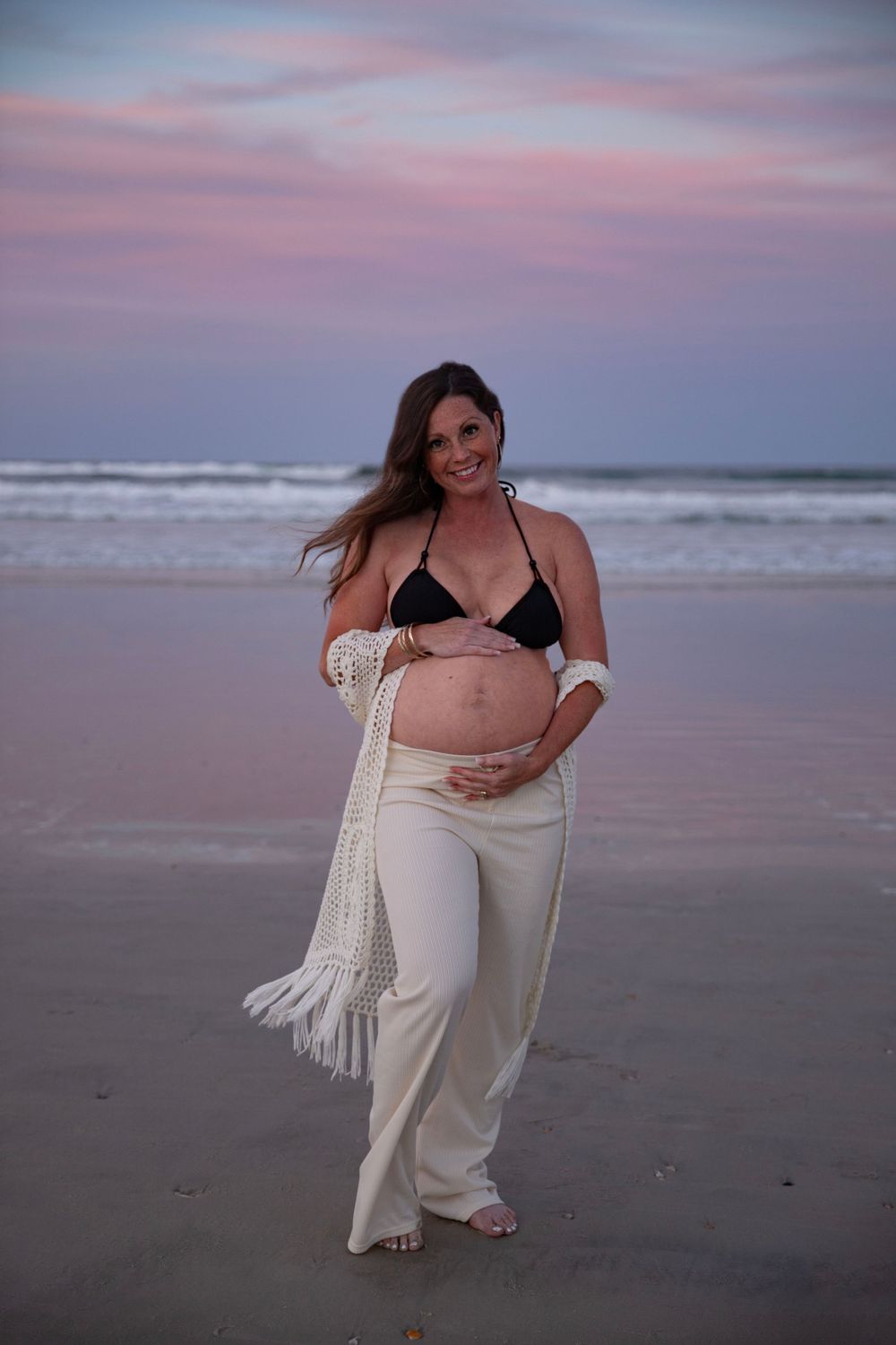 Pregnant woman in black bikini top and white pants holding baby bump on beach at sunset.