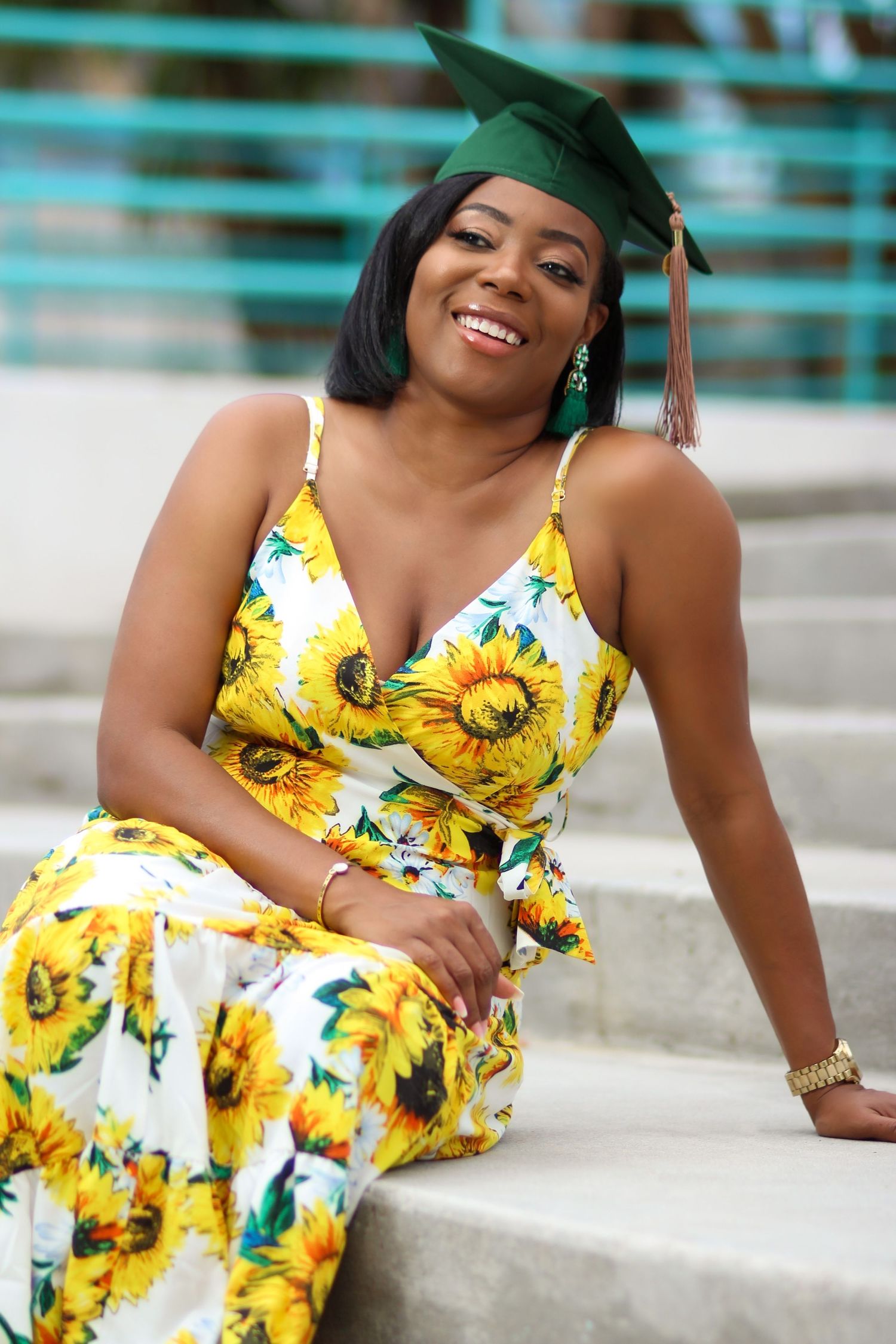 Graduate in sunflower print dress poses on concrete steps wearing academic cap.
