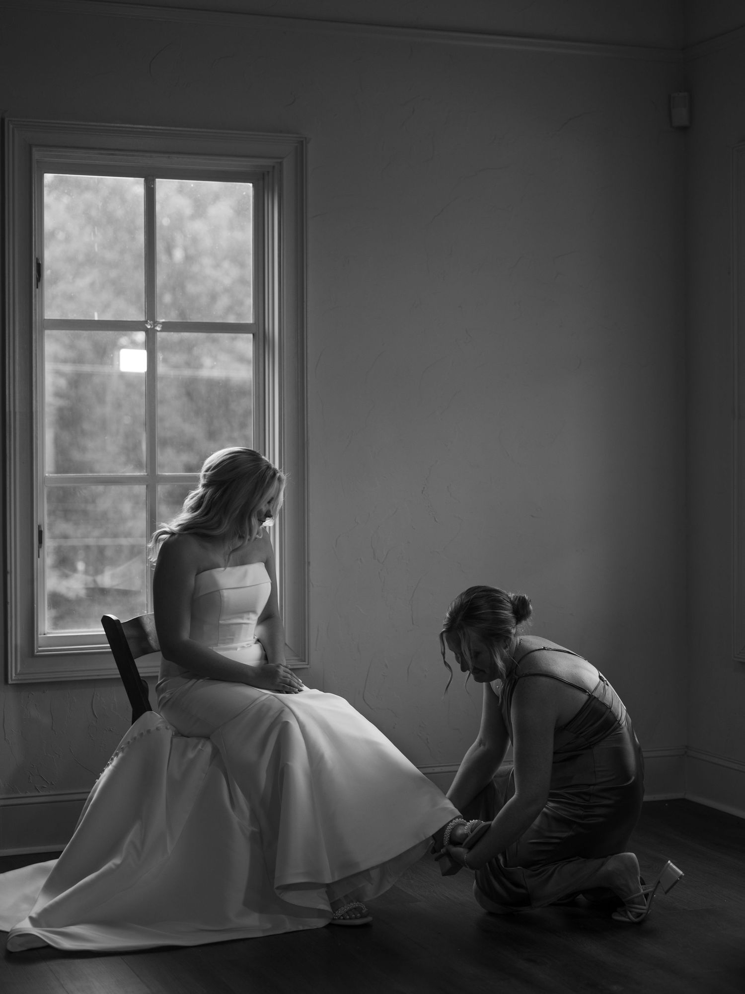 Black and white photo of wedding preparations as a bride in white dress sits by window while helper adjusts the gown.