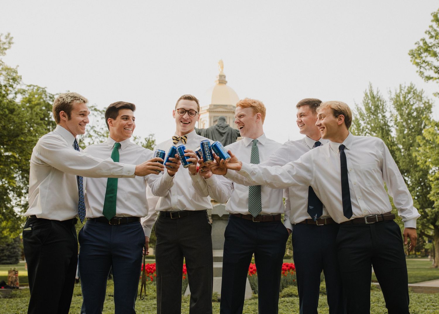 Group of groomsmen toasting with bottles on grass with dome in background.
