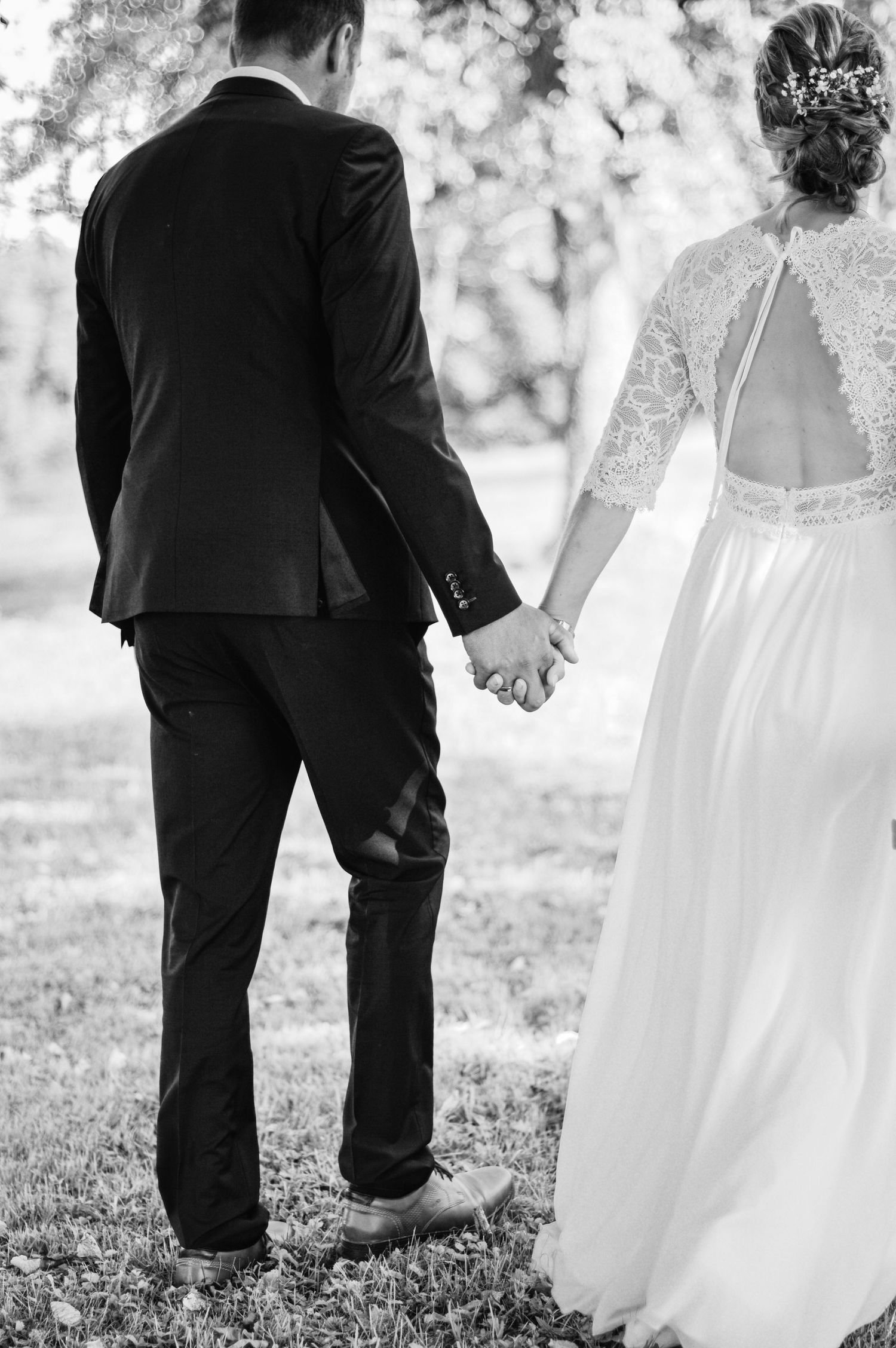 Schwarz-Weiß-Hochzeitsfoto, das die Braut im Spitzenkleid und den Bräutigam im Anzug zeigt, die sich in einem Garten im Freien an den Händen