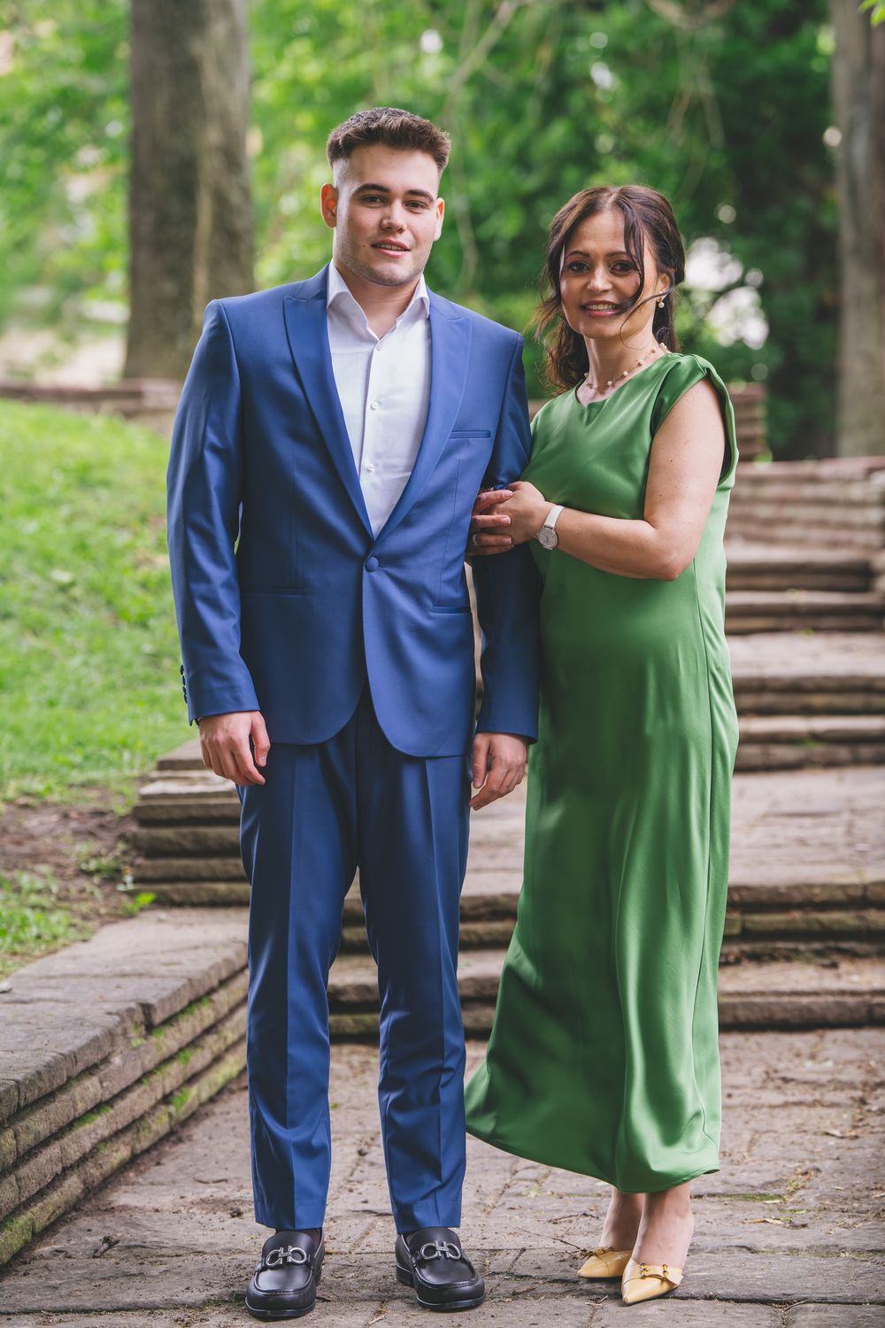 A pair in formal attire poses on stone steps in a park, with one wearing a blue suit and another in a green dress.