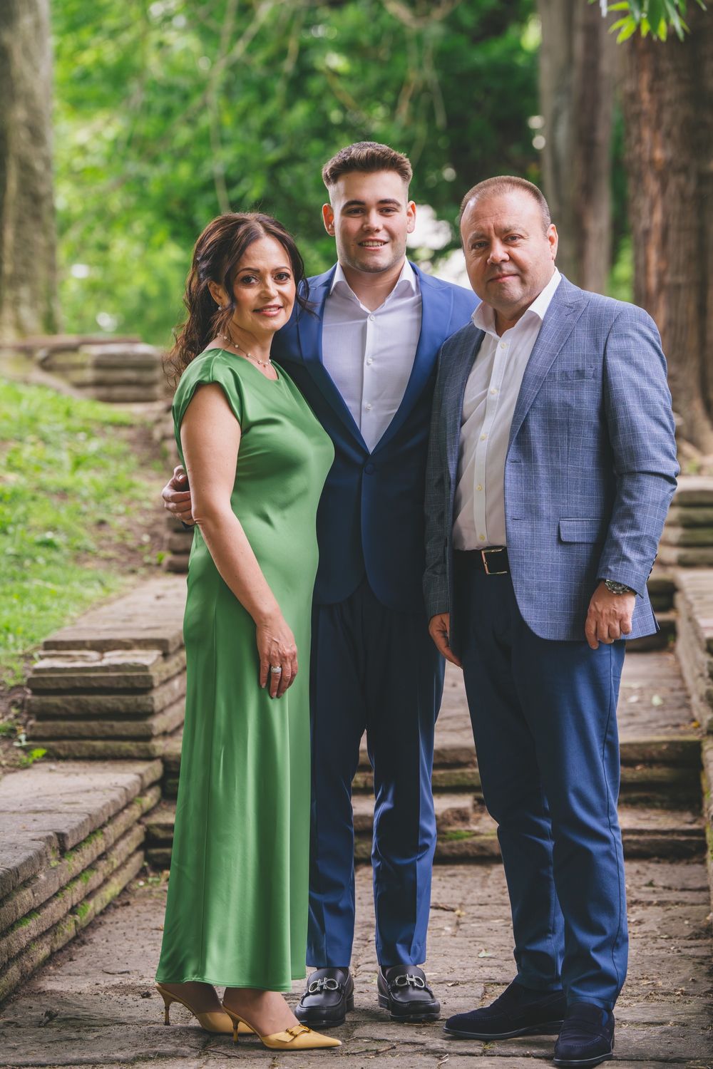 Three people in formal wear pose together on outdoor stone steps surrounded by greenery.