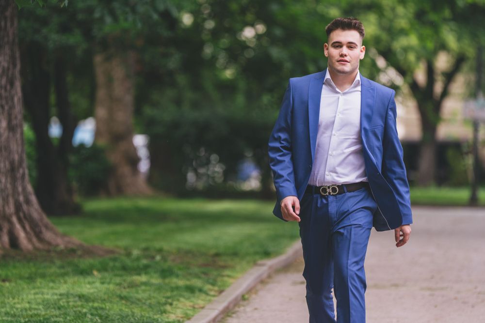 A person in a blue suit and white shirt walks confidently along a tree-lined path.