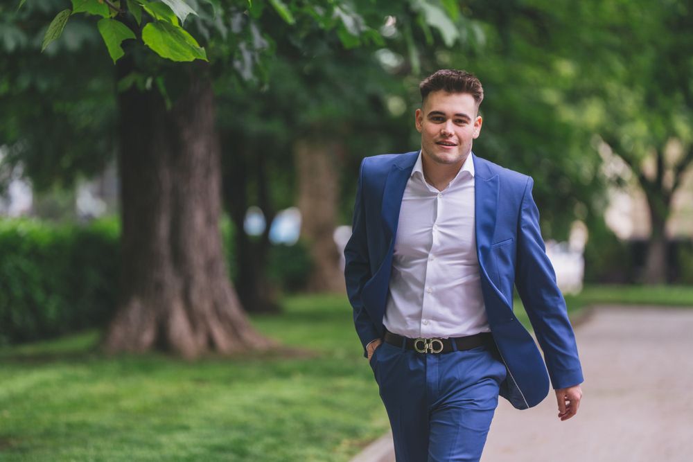 A person strolls through a park wearing a tailored blue suit with white shirt.