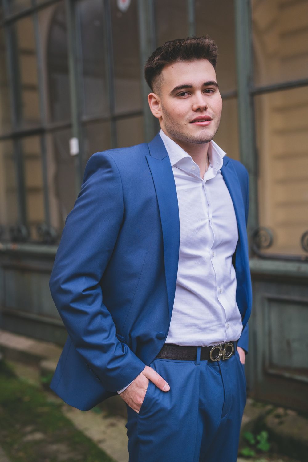 A person in a fitted blue suit and white dress shirt poses outdoors near a building.