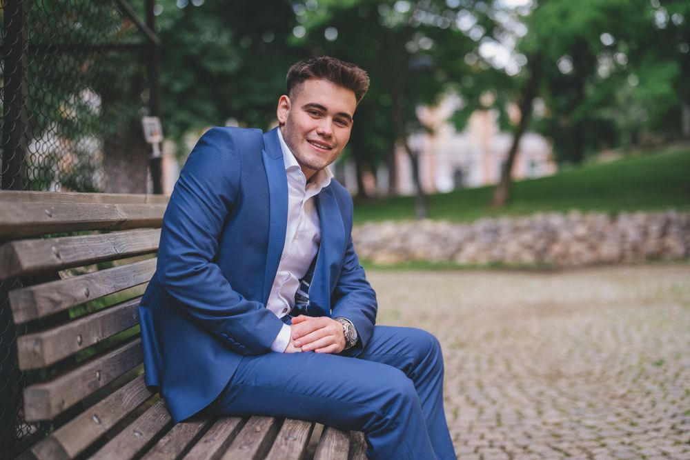 A person in a blue suit sits casually on a wooden bench in an outdoor setting.