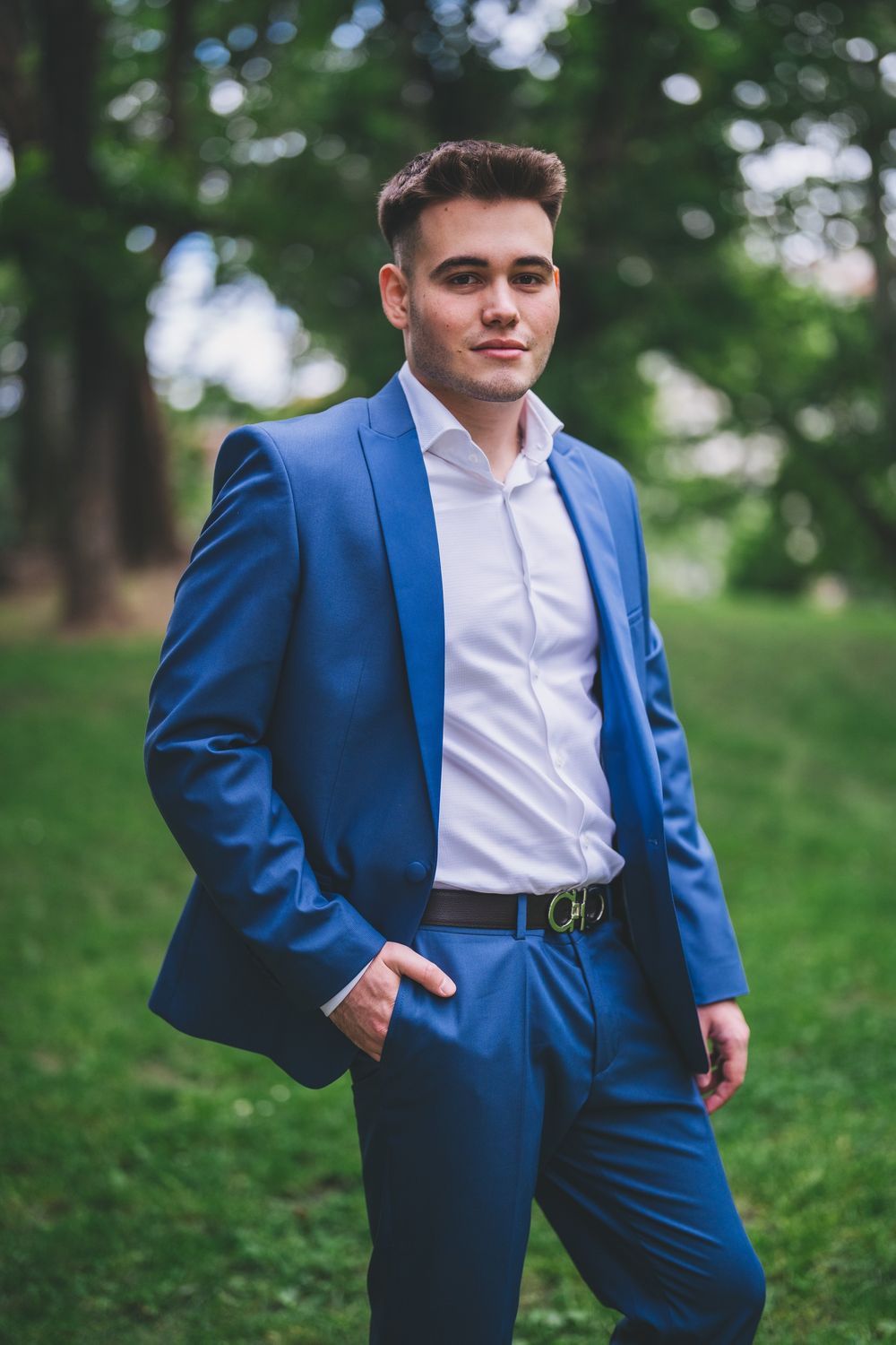 A person in a tailored blue suit poses confidently in a lush green park setting.