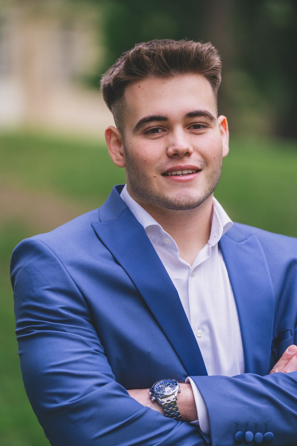 Professional portrait of someone in a blue blazer and white dress shirt smiling outdoors with greenery in background.