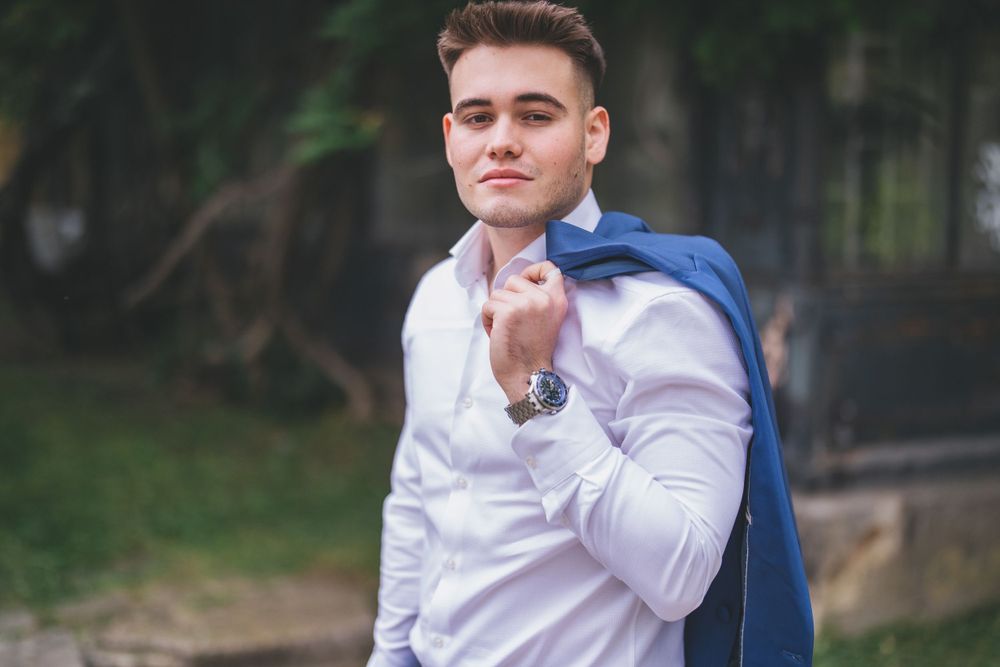 Person in business attire poses casually with a blue blazer draped over their shoulder in a garden setting.