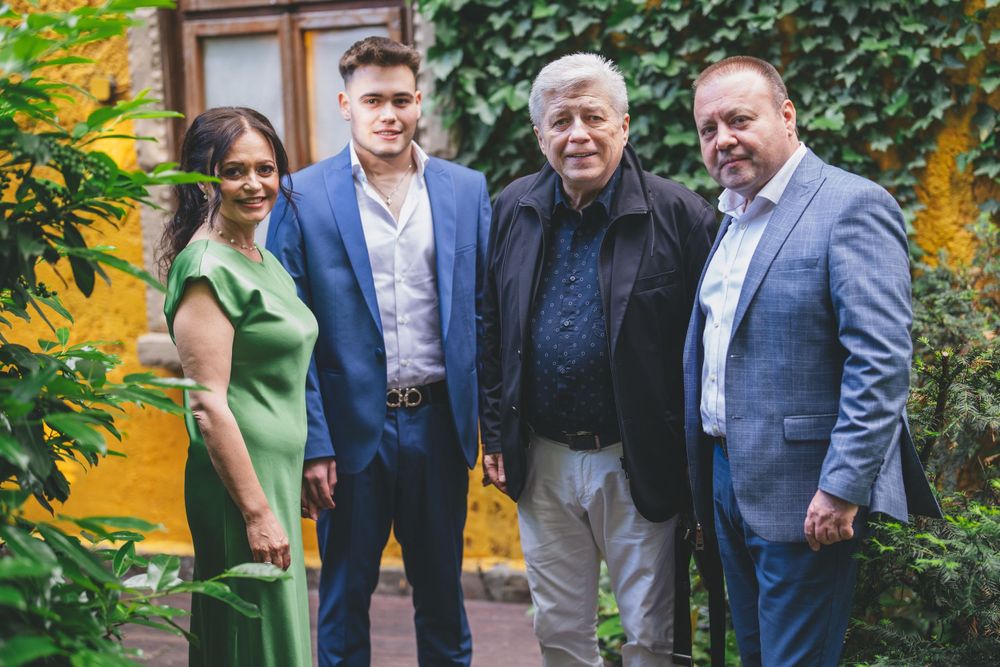 Group of people dressed formally pose together outdoors against a yellow wall with greenery.